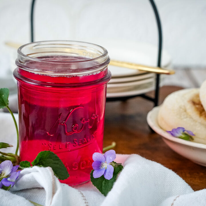 A jar of wild violet jelly with several violets to one side and a plate of English muffins beside.