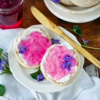 A white plate with two English muffins spread with violet jelly and garnished with violet flowers.
